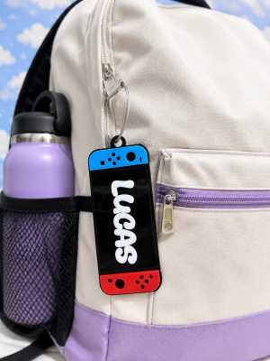 Personalized acrylic game console backpack tag attached to a school backpack with a custom name.